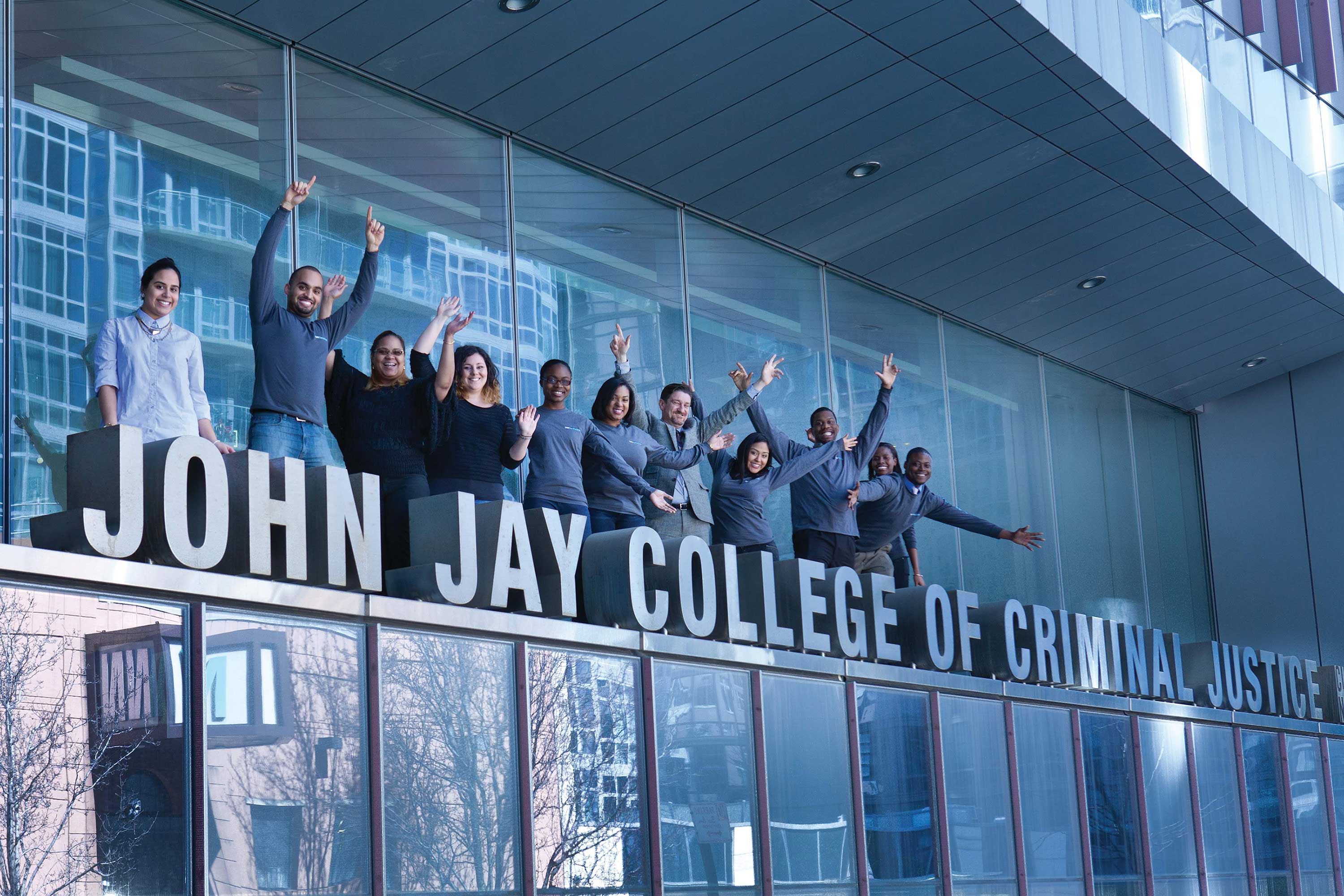 A group of diverse students and staff stands behind the large "JOHN JAY COLLEGE OF CRIMINAL JUSTICE" sign, cheering with their arms raised.