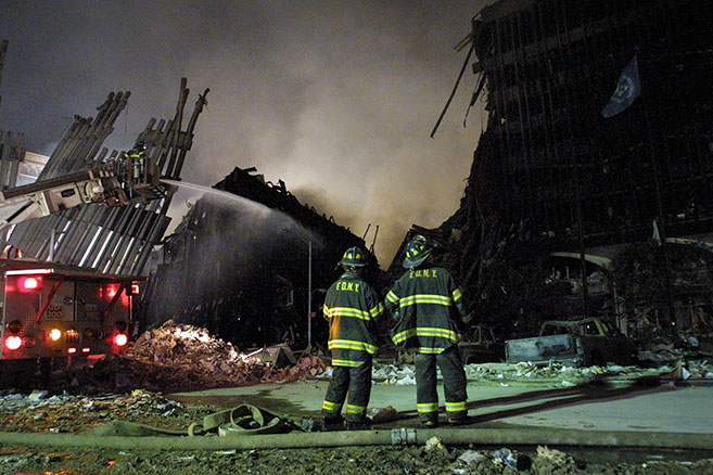 Two firefighters staring at buildings that have been burned down with a firetruck spraying water on the debris