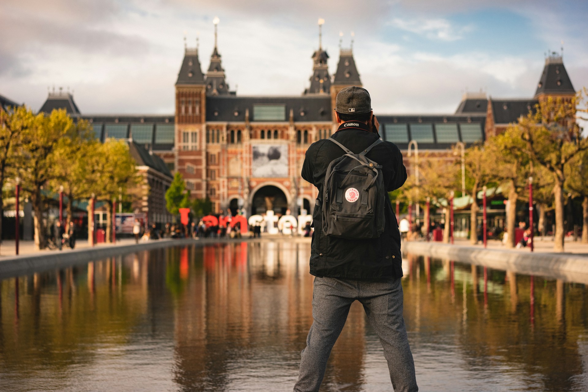 Male wearing backpack facing away from camera taking photo of a river and brick building