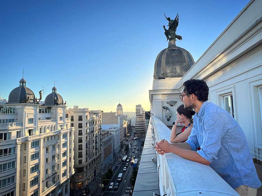 Two people on a balcony overlooking a grand Madrid street with ornate domed buildings and statues.