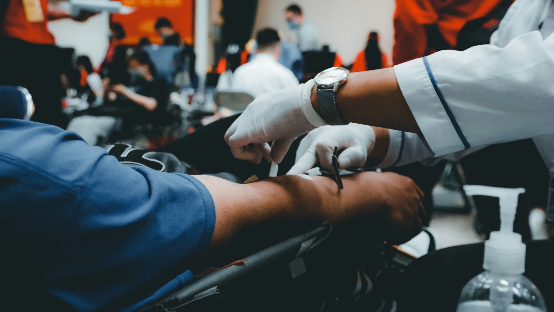 Close-up of a healthcare worker wearing gloves inserting a needle into a person’s arm during a blood donation, with other donors and staff blurred in the background.