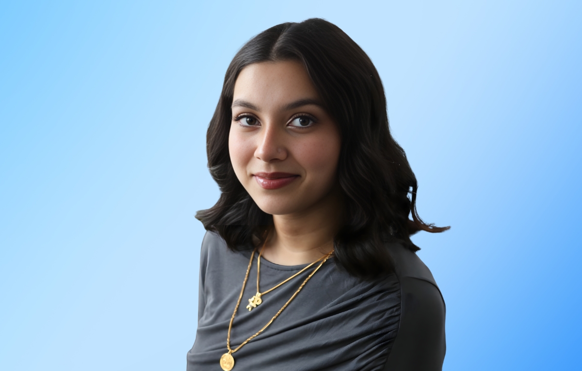 Ayesha Akter, smiles and faces the camera. Her hair is down, parted in the middle. She is wearing a grey top and two gold necklaces.