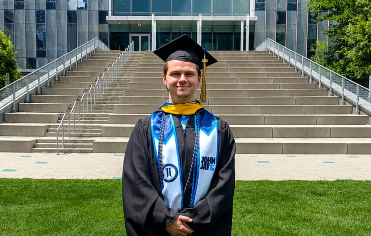 Luke Mullane stands in front of the stairs on the Jay Walk. He is wearing his graduation cap and gown, his John Jay Stole, and is smiling at the camera. 
