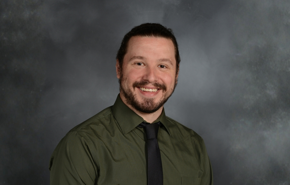 Richard Piszczatowski sits smiling at the camera. He's wearing a deep hunter green button down and black tie. The background is a dark gray.