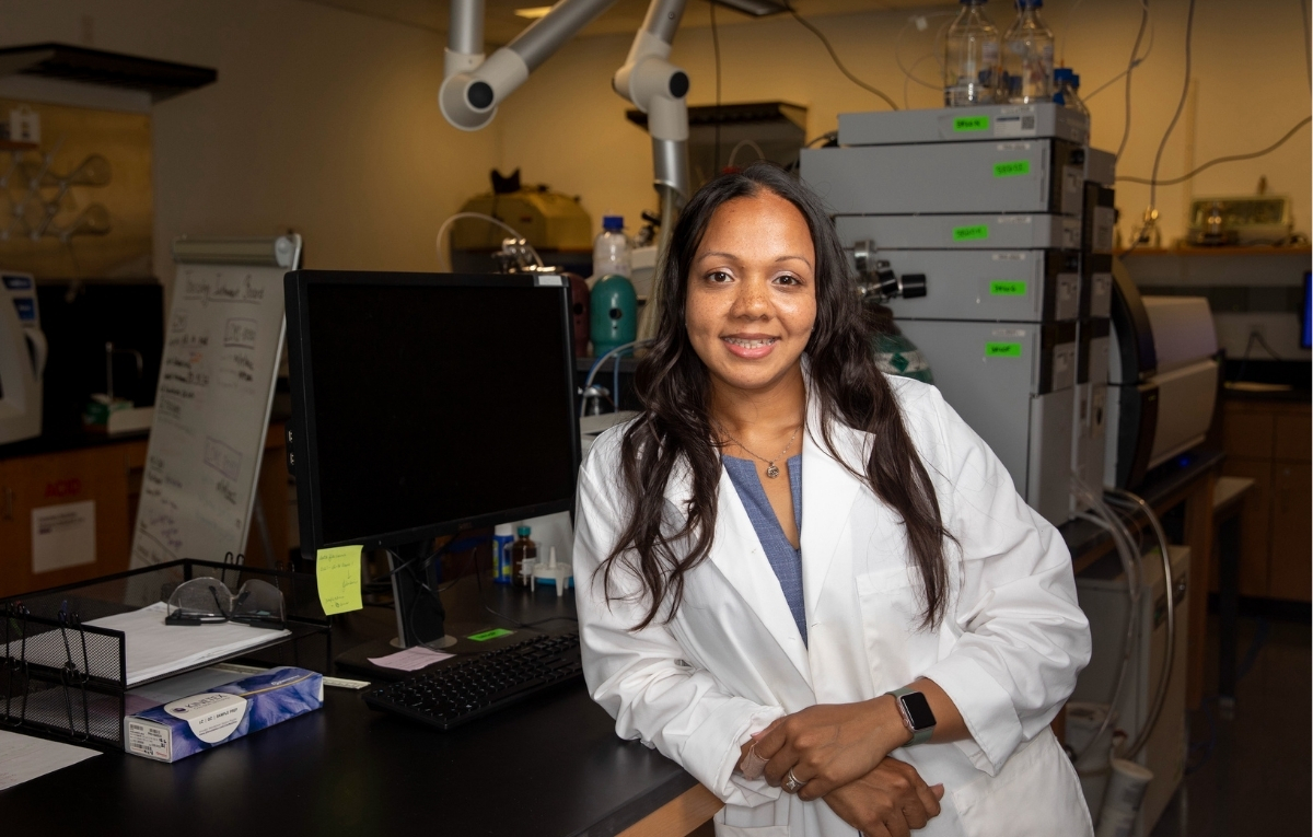 Teeshavi Acosta wears a lab coat and leans up against one of the tables in her science lab. She is smiling. 