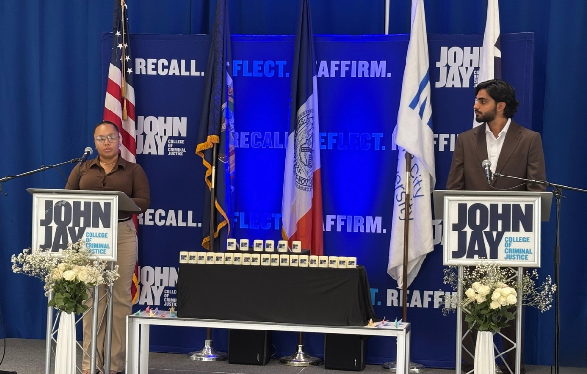Two students (Dane Cotes on left and Ali Hassan on right) stand at podiums during a 9/11 remembrance event at John Jay College, with American and college flags and a table of memorial candles between them.