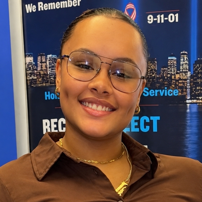 Portrait of a smiling student, Dane Cotes, wearing glasses and a brown shirt, standing in front of a 9/11 remembrance display.