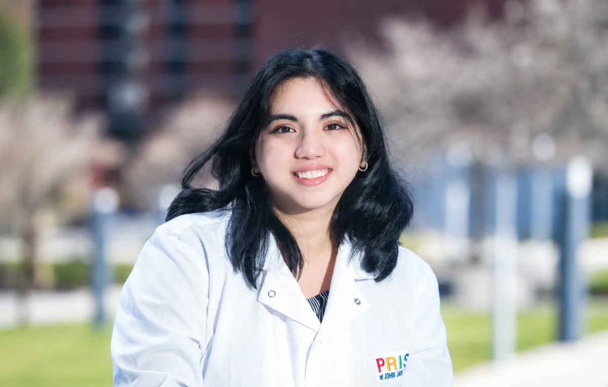 A smiling alumna, Maisha Ahmed, wears a white lab coat embroidered with the John Jay College PRISM logo poses outdoors on the Jay Walk.