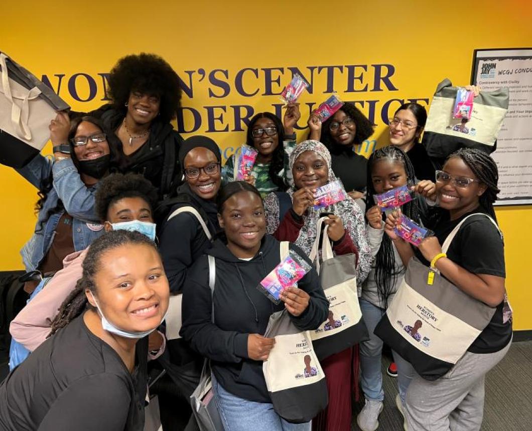 A group of female students posing in front of a yellow wall that says Women's Center for Gender Justice. They're all showing off their similar totebags and goodies from an event.