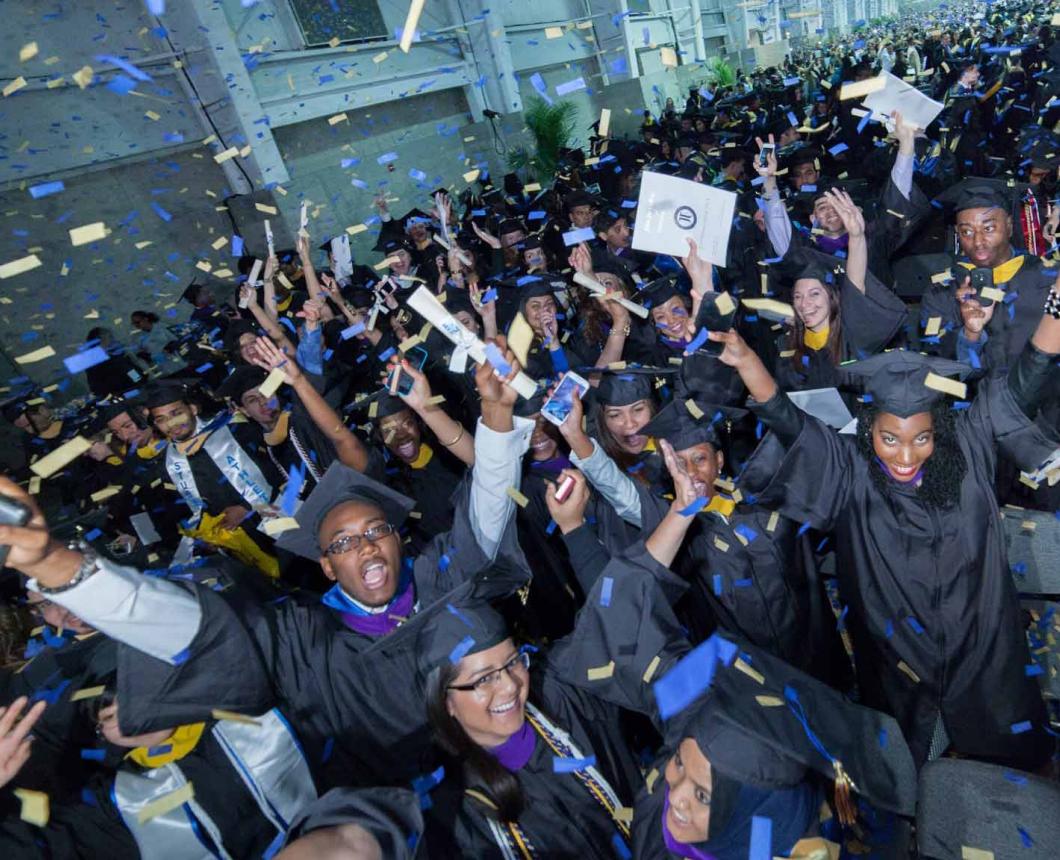 A large crowd of John Jay College graduates in caps and gowns cheer with their arms raised as gold and white confetti rains down at a commencement ceremony.
