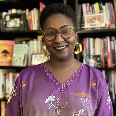 Head shot of professor Moffett-Bateau. She has a short afro with the sides cut short. They are wearing gold hoop earrings, gold large glasses and a purple dashiki.