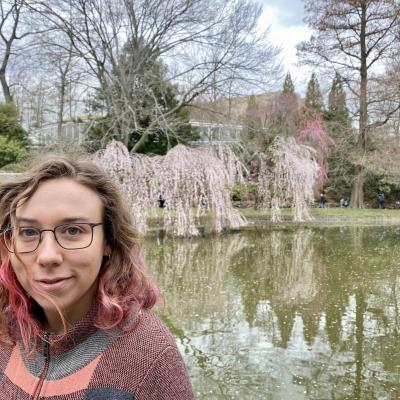 a woman with glasses and brown hair with pink streaks stands in front of a lake and flowering trees